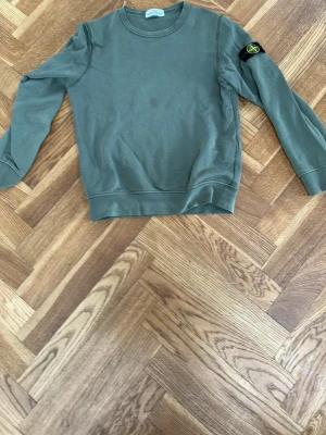 Grön sweatshirt från Stone Island - Säljer en grön sweatshirt från Stone Island med klassisk rund hals och ribbade muddar. Tröjan har den ikoniska Stone Island-loggan på vänster ärm och är tillverkad i mjuk bomull. Perfekt för en avslappnad och stilren look.