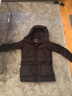 Svart lång dunjacka med huva - Snygg svart dunjacka med huva och långa ärmar. Jackan har dragkedja och knappar framtill, två stora fickor med lock och två bröstfickor. Lång modell som håller dig varm under kalla dagar. Perfekt för vintern! Har småa hål 