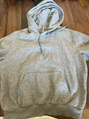 Grå hoodie med magficka - En klassisk grå hoodie med huva och dragsko. Hoodien har en stor magficka framtill och ribbade muddar vid ärmslut och nederkant. Perfekt för en avslappnad och stilren look.