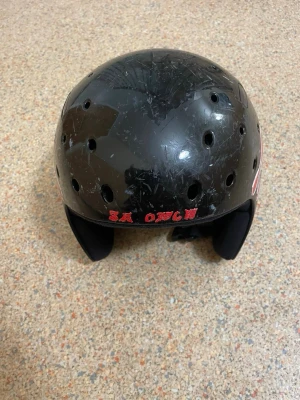 Svart hjälm med ventilationshål - Svart hjälm med flera ventilationshål och vadderade öronskydd. Utrustad med justerbart hakband och text i rött framtill. Passar till exempelvis skateboard, cykel eller annan sport. Tillverkad i hårdplast med mjuk insida.