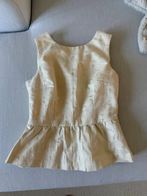 Beige ärmlös topp med dragkedja - Snygg beige ärmlös topp med subtilt mönster och peplumvolang. Toppen har en v-ringning och dragkedja bak för en cool detalj. Perfekt att styla med jeans eller kjol för en trendig look.