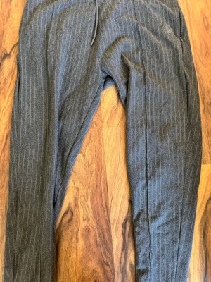 Gråa mjukisbyxor från SikSilk - Mörkgrå mjukisbyxor från SikSilk med tunna vita ränder och dragsko i midjan. Byxorna har en avslappnad passform och är perfekta för en chill stil. Materialet känns mjukt och bekvämt, och loggan är diskret broderad på ena benet.