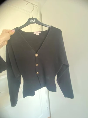 Svart kofta med guldknappar - Stilren svart kofta från Gina Tricot med djup V-ringning och tre dekorativa guldknappar framtill. Modellen har lång ärm och är tillverkad i mjukt, tunt stickat material. Perfekt att slänga över en topp för en enkel men snygg look.