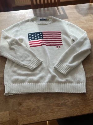 Vit stickad tröja Ralph Lauren - Vit stickad tröja från Ralph Lauren med amerikansk flagga broderad på bröstet och RL-bokstäver. Tröjan har rund hals, långa ärmar och ribbstickade muddar samt nederkant. Perfekt för dig som gillar klassisk och stilren design.