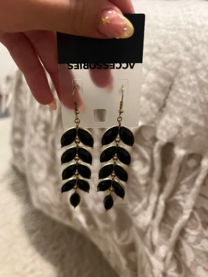 Svarta bladformade örhängen Accessorize - Snygga örhängen från Accessorize med bladformade hängen i svart och guldfärgade detaljer. Örhängena har en elegant design med flera små blad i rad och krokfäste. Perfekta för att lyfta din outfit med en trendig touch.