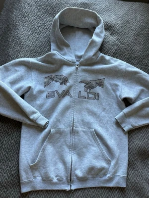 Grå hoodie med tryck från BVALDI - Snygg grå zip-hoodie från BVALDI med tryck av två händer och text på bröstet. Hoodien har huva, dragkedja framtill och en stor magficka. Perfekt för en avslappnad och cool stil.