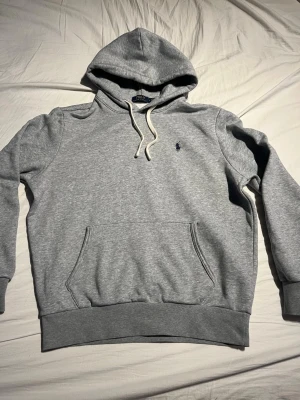 Grå hoodie från Polo Ralph Lauren - Snygg grå hoodie från Polo Ralph Lauren med klassisk blå logga på bröstet. Hoodien har vit dragsko i huvan och en stor magficka framtill. Tillverkad i mjukt bomullsmaterial och har ribbade muddar vid ärmslut och nederkant.