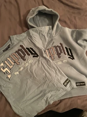 Blå t-shirt från Supply - Blå t-shirt från Supply med stor tryckt logga i svart och beige framtill. Klassisk rund hals och korta ärmar. T-shirten har en modern streetwear-look och är tillverkad i mjuk bomull för skön känsla.