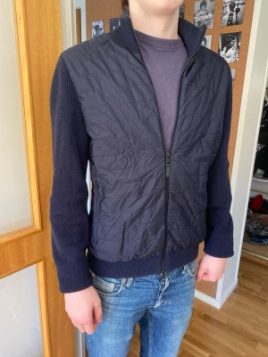 Mörkblå cardiganjacka med quiltad front - Snygg mörkblå cardiganjacka med quiltad front och stickade ärmar. Jackan har dragkedja hela vägen, hög krage och ribbade muddar. Perfekt för lager på lager och ger en modern look till din outfit.