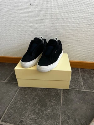 Svarta sneakers från Axel Arigato - Snygga svarta sneakers från Axel Arigato i mocka med vit tjock sula. Diskret logga i guld på sidan och stilren slip-on design utan snörning. Perfekta för dig som gillar minimalistisk och clean stil. JAG HAR ANVÄNT DOM FYRA GÅNGER BARA!!! HAR DU NÅGRA FRÅGOR ÄR DET BARA ATT SKRIVA TILL MIG!!! VI KAN DISKUTERA OM PRISET!!!!