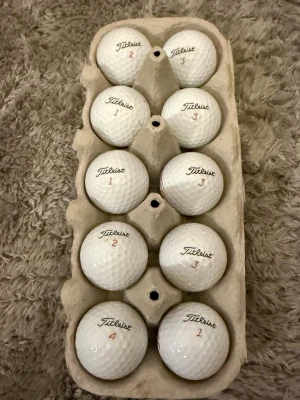 Titleist vita golfbollar TruFeel a-b klass - Säljer ett set med vita golfbollar från Titleist, modell TruFeel. Bollarna har klassisk dimple-design och är märkta med siffror och röd text. Perfekta för dig som vill ha kvalitetsbollar till golfbanan.