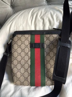 Gucci axelväska med GG-mönster - Äkta köpt hos circular fashion har äktehets intyg Snygg axelväska från Gucci i beige canvas med klassiskt GG-mönster och bred grön-röd rand framtill. Väskan har svarta läderdetaljer och justerbar axelrem i textil med läderförstärkning. Perfekt för dig som gillar lyxiga accessoarer med ikonisk design.