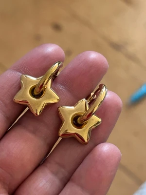 Guldiga stål örhängen med stjärnhänge - Snygga örhängen i stål guldfärg med chunky stjärnformade hängen. Örhängena har en rund ring som håller i en glansig stjärna, vilket ger en cool och trendig look. Perfekta för dig som gillar statement-smycken och vill sticka ut lite extra.