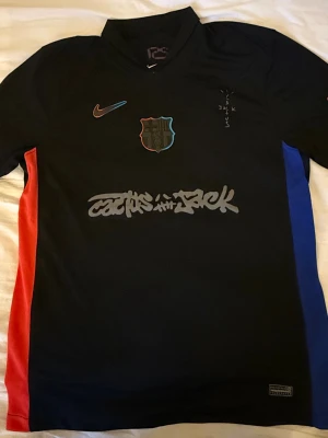 FC Barcelona x Nike svart matchtröja - Svart FC Barcelona matchtröja från Nike med färgdetaljer i rött och blått på sidorna. Framsidan har klubbmärke, Nike-logga och grafiskt tryck. Baksidan har UNHCR-logga och gula ränder i nacken. Perfekt för dig som älskar fotboll och streetstyle.