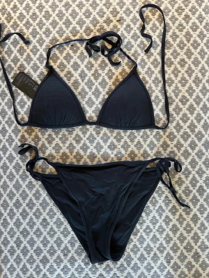 Svart bikini med knyt från H&M  - Snygg svart bikini från hm  med klassisk trekantsform och knyt både i nacken och på sidorna av bikinitrosan. Fodrade kupor . Stl 34 . Använd en gång 