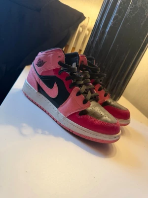 Air Jordan 1 Mid Pink/Black - Snygga Air Jordan 1 Mid sneakers i rosa, svart och vitt läder. Klassisk siluett med svart snörning, rosa swoosh och detaljer samt perforerad tå. Perfekta för dig som vill sticka ut med färg och ikonisk design. Skickas med ljusrosa snören till.