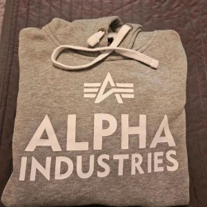 Grå hoodie från Alpha Industries - Grå hoodie Alpha Industries S Snygg grå hoodie från Alpha Industries med stor vit logotyp på bröstet och klassisk huva med snörning. Mjuk insida och ribbade muddar vid ärmar och nederkant. Perfekt för chill dagar och streetstyle.  Kan även hämtas I Partille  E1