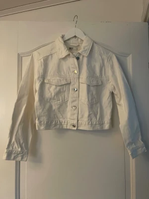 Vit jeansjacka - Snygg vit jeansjacka med klassisk krage. Jackan har två bröstfickor med lock och silverfärgade knappar framtill. Croppad modell som passar perfekt till både jeans och kjol. Långärmad och i ett stadigt jeanstyg.