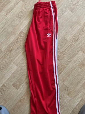 Röda Adidas mjukisbyxor med vita ränder - Snygga röda mjukisbyxor från Adidas med klassiska vita ränder längs sidorna och Adidas-logga på låret. Byxorna har resår i midjan och dragkedja på fickan. Tillverkade i ett mjukt och följsamt material som passar perfekt för en avslappnad stil.