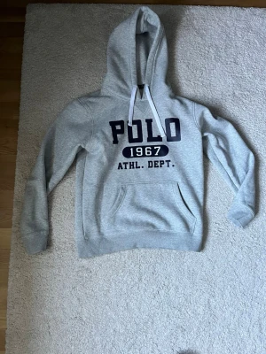 Grå hoodie från Polo Ralph Lauren - Snygg grå hoodie från Polo Ralph Lauren med stor svart text 'POLO 1967 ATHL. DEPT.' på bröstet. Klassisk känguruficka, vita dragsnören och mjukt material. Perfekt för en avslappnad och sportig stil.