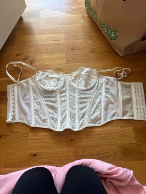 Vit spetskorsett med bygel - Säljer en vit korsett med bygel och spetsdetaljer. Den har justerbara axelband, hakknäppning bak och vackra broderade partier framtill. Materialet är en mix av spets och mesh som ger en elegant och feminin look.