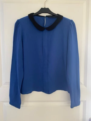 Blå blus med svart krage från Zara - Snygg blå blus från Zara Woman med lång ärm och kontrasterande svart rund krage. Tillverkad i ett lätt, mjukt tyg som ger ett stilrent intryck. Perfekt för dig som gillar klassiska detaljer och enkel design.