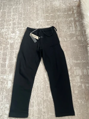 Svarta mjukisbyxor Essentials - Svarta mjukisbyxor från Fear of God Essentials med dragsko i midjan och elastisk resår. Byxorna har en stilren design med diskret logga framtill och är tillverkade i mjukt material som känns skönt mot huden. Perfekta för en avslappnad och trendig look.