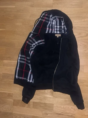 Svart hoodie från Burberry - Svart hoodie från Burberry med dragkedja och klassiskt rutigt foder i huvan. Hoodien har ribbade muddar och en stilren look med diskret logga på bröstet. Perfekt för dig vill ha något som sticker ut men ändå med kavlite.