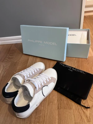 Philippe Model vita sneakers med beige detaljer - Snygga vita sneakers från Philippe Model med beige mockadetaljer och svart häl. Klassisk låg modell med vita skosnören och diskret logga på sidan. Skorna har en stilren design och är tillverkade i läder och mocka.