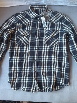 Svartvit flanell rutmönstrad skjorta Diesel - Säljer en oanvänd svartvit rutmönstrad skjorta från Diesel med klassisk krage och två bröstfickor med lock. Skjortan har långa ärmar och knäppning framtill. Perfekt för dig som gillar en avslappnad och trendig look.