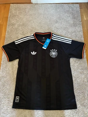 Svart Adidas Tyskland matchtröja - Snygg svart matchtröja från Adidas med Tysklands landslagsmärke och fyra stjärnor på bröstet. Tröjan har vita ränder på axlarna, diskreta mönster i tyget och orange detaljer vid ärmslut och krage. Tillverkad i lätt och ventilerande polyester.