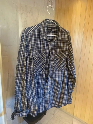 Blårutig skjorta från Tuxer - Snygg blårutig skjorta från Tuxer med klassisk krage och två bröstfickor med knappar. Skjortan har långa ärmar och är tillverkad i ett mjukt bomullsmaterial. Perfekt för en avslappnad och cool stil. Skicket är i utmärkt skick och hör bara av dig om du har några vidare funderingar🙌