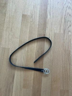 Svart smalt bälte med silverring - Stilrent svart bälte i skinn med smal design och dubbla silverfärgade ringar som spänne. Perfekt accessoar för att lyfta din outfit med en enkel men trendig detalj.