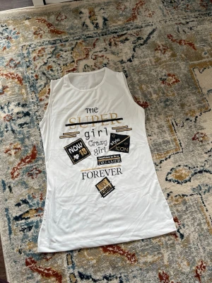 Vit ärmlös linnetopp med tryck - Vit ärmlös linnetopp med tryck i svart och guld. Texten på framsidan har detaljer som 'SUPER girl', 'style ICON' och 'FOREVER'. Toppen har rund halsringning och är tillverkad i ett mjukt, lätt material.