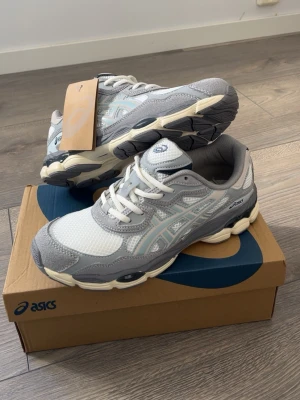 ASICS grå och vita sneakers - Snygga ASICS sneakers i grått och vitt med mesh och syntetmaterial. Skorna har klassisk låg profil, vita snören och detaljer i ljusblått. Sulan är chunky och har beige inslag för en modern look. Perfekta för dig som gillar sportig stil.