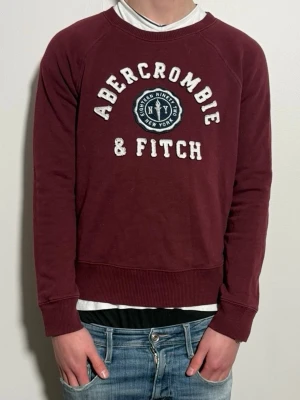 Vinröd sweatshirt Abercrombie & Fitch - Snygg vinröd sweatshirt från Abercrombie & Fitch i storlek S. Klassisk modell med broderad logga framtill. Bra skick, inga större defekter. Perfekt till vardag. Modellen är 176 cm och väger 65kg då den är för lite. OBS ÄR UNISEX.