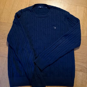 Mörkblå kabelstickad tröja från GANT - Säljer en mörkblå kabelstickad tröja från GANT med rund hals och diskret logotyp broderad på bröstet. Tröjan har långa ärmar och ribbade muddar vid ärmslut och nederkant. Perfekt för dig som gillar klassisk och stilren design. Passar 170-176cl