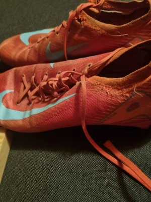 Nike röda fotbollsskor med blå detaljer - Säljer ett par röda Nike fotbollsskor med ljusblå swoosh och detaljer. Skorna har snörning och en smal, strömlinjeformad design med plastdobbar på sulan för bra grepp på planen. Materialet är syntetiskt och lätt för optimal prestation.
