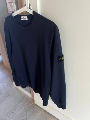 Stone Island tröja 170 - Skick 8/10. Strl 170. Mörkblå sweatshirt från Stone Island Junior med klassisk rund halsringning och ikonisk logotyp-patch på vänster ärm. Tröjan har ribbade muddar vid ärmslut och nederkant. Perfekt för en stilren och avslappnad look. 