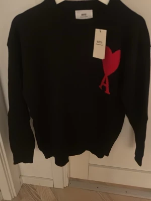 Svart stickad tröja från Ami Paris - Cool svart stickad tröja från Ami Paris med röd hjärtlogo och bokstaven A på bröstet. Tröjan har rund hals och långa ärmar, perfekt för att ge din outfit en trendig touch. Materialet är mjukt och skönt, idealiskt för lager-på-lager.