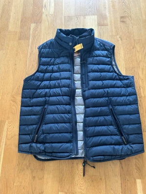 ÄKTA! Mörkblå dunväst från Parajumpers - L men sitter som M                                         Snygg mörkblå dunväst från Parajumpers med quiltad design och hög krage. Västen har två dragkedjefickor framtill och en ikonisk gul detalj vid halsen. Insidan är ljusgrå och västen stängs med dragkedja. Perfekt för lager-på-lager-stil.
