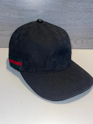 Svart keps från Gucci med logomönster - Snygg svart keps från Gucci med diskret GG-logomönster i tyget. Kepsen har klassisk böjd skärm och detaljer med grönt och rött band på sidorna. Tillverkad i textilmaterial som ger en exklusiv känsla och passar perfekt till streetwear-stilen.