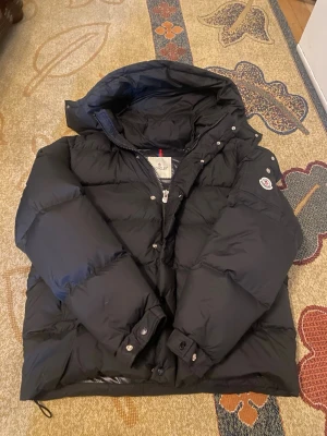 Svart Moncler dunjacka med hög krage - Säljer en svart dunjacka från Moncler med hög krage och knappar framtill. Jackan har två stora fickor med dragkedja och snörning nedtill. Klassisk Moncler-logga på ärmen. Perfekt för kalla dagar, riktigt varm och stilren.