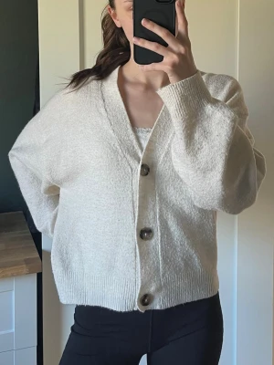 Beige oversized kofta med knappar - Mysig beige oversized kofta med stora bruna knappar framtill och ribbade muddar. Koftan har v-ringning och är gjord i ett mjukt, stickat material som känns skönt mot huden. Perfekt för lager på lager och en avslappnad look.