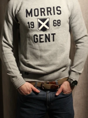 Grå tröja Morris Gent 1968 - Snygg grå tröja från Morris med trycket 'MORRIS GENT 1968' och ett skotskinspirerat emblem på bröstet. Tröjan har rund hals, ribbade muddar och är tillverkad i mjukt stickat material. Perfekt för dig som gillar klassisk men sportig stil.