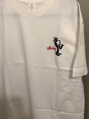 Vit Stüssy t-shirt med grafiskt tryck - Vit t-shirt från Stüssy med coolt grafiskt tryck på både fram- och baksidan. Framsidan har en liten svart figur och Stüssy-logga i rött, medan baksidan har ett stort konstnärligt motiv med text runtom. Klassisk passform och rund hals.