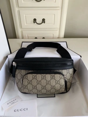 Beige midjaväska med monogram - Snygg shoppingväska i beige canvas med klassiskt monogram och svarta detaljer i läder. Väskan har en justerbar svart axelrem och en praktisk framficka med dragkedja. Perfekt för dig som vill ha en trendig och lyxig accessoar.