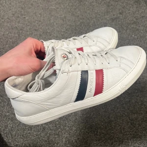 Vita Moncler sneakers med röd/blå detalj - Snygga vita sneakers från Moncler i läder med klassiska röda och blå ränder på sidan samt Moncler-logga på plösen. Skorna har vita snören och en låg silhuett som ger en clean och stilren look. Perfekta för dig som gillar exklusiva och tidlösa sneakers. Storlek 43, men sitter mer som 44! 
