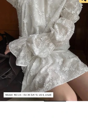 Vit blommig kortklänning med spets - Superfin vit kortklänning med broderade blommor och spetsdetaljer. Klänningen har långa puffärmar, öppen rygg med knyt och volang nertill. Perfekt för sommarens alla tillfällen! Från Nakd. Storlek 34 och använd en gång! Perfekt till student eller konfirmation 