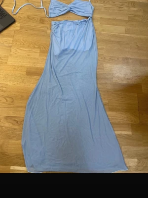 Ljusblått tvådelat set med twisttopp - Säljer ett ljusblått tvådelat set med en twistad crop top med smala axelband och en lång, mjuk kjol med resår i midjan. Setet är gjort i ett tunt och stretchigt material som känns bekvämt mot huden. Perfekt för dig som gillar en trendig och avslappnad look.
